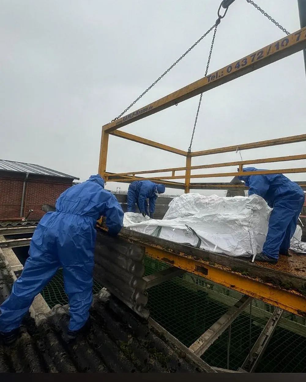 Drei Mitarbeiter in blauen Einwegschutzanzügen heben verpackte Asbestzementplatten von einem Welleternitdach auf eine Kranbühne.