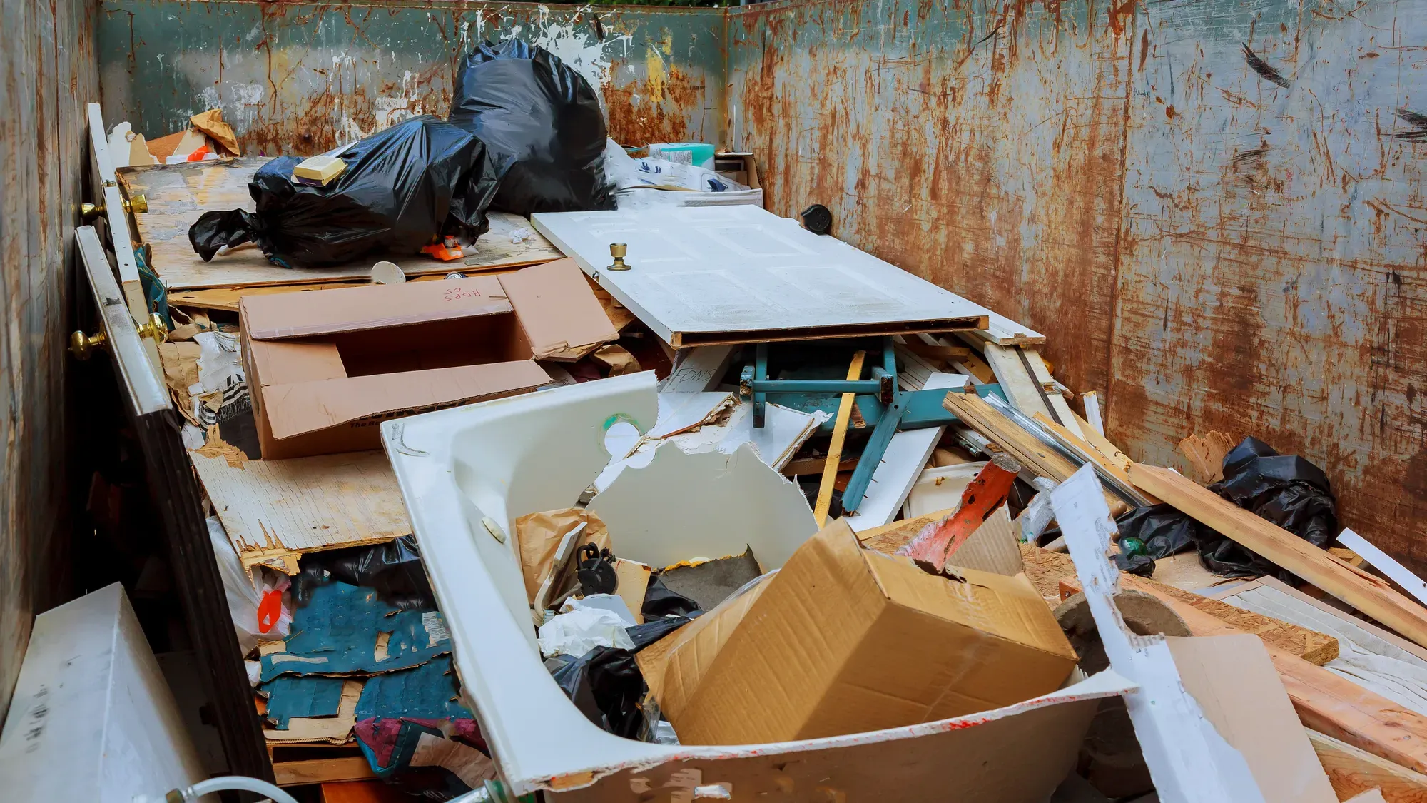 Blick in einen gefüllten Abrollcontainer einer Räumung: eine ausgehängte Zimmertür, eine ausgebaute Badewanne, Kartonagen, Holzreste und verschnürte Müllsäcke.