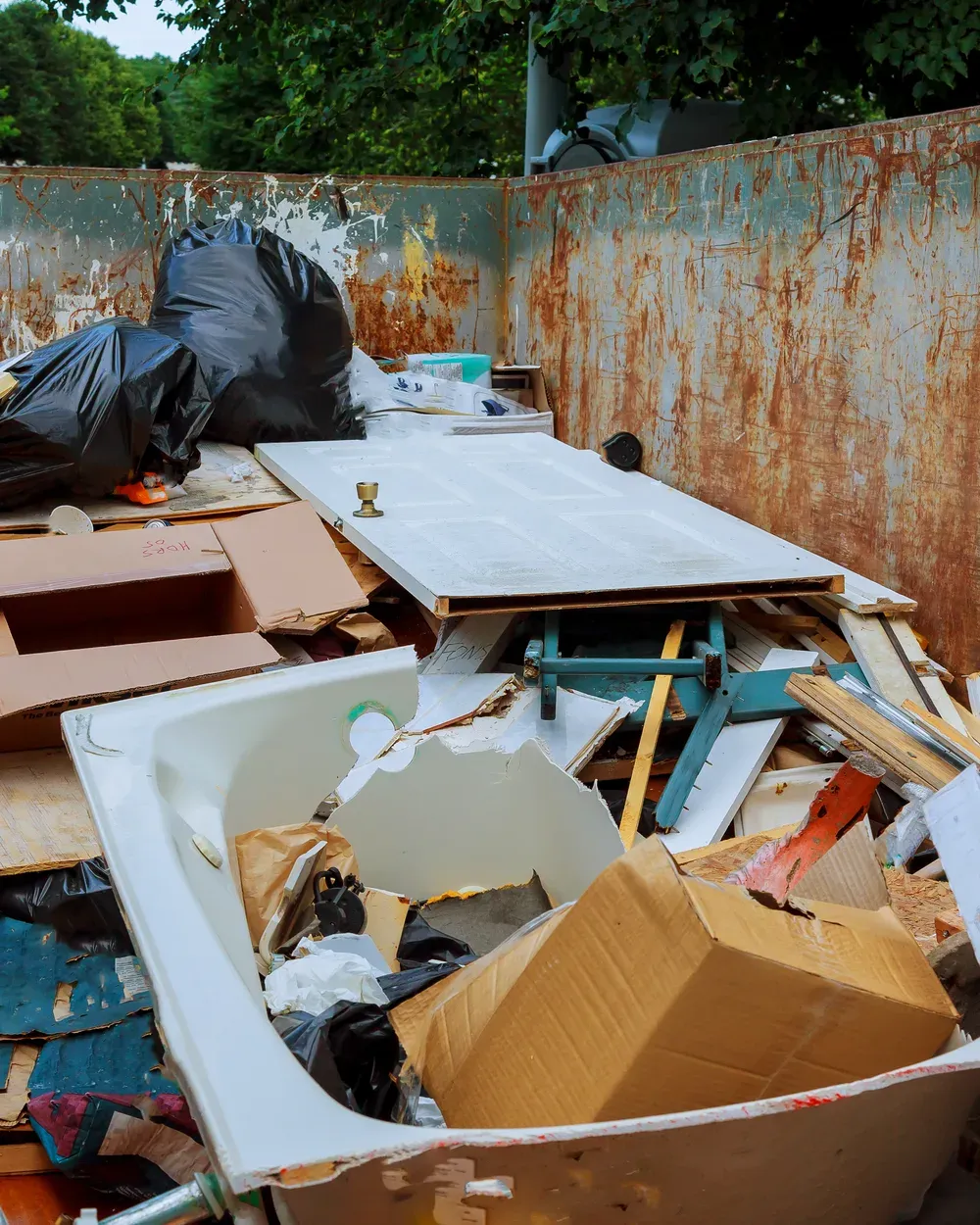 Hochformatiger Blick in denselben Container: Badewanne, Zimmertür, ein zerlegter Hocker, Kartonagen und Bauholz liegen übereinander.