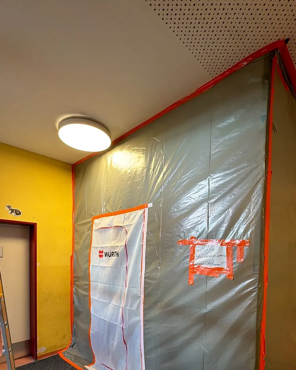 Mit Folie staubdicht abgeschotteter Arbeitsbereich in einem Flur: Die Ränder sind mit orangefarbenem Klebeband verklebt, der Zugang führt durch eine Staubschutztür mit Reißverschluss.