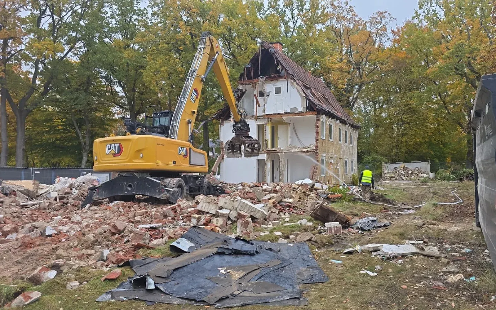 Kettenbagger mit Abbruchgreifer trägt ein zweigeschossiges Wohnhaus ab; im Vordergrund liegt sortierter Ziegelbruch, ringsum herbstliche Laubbäume.